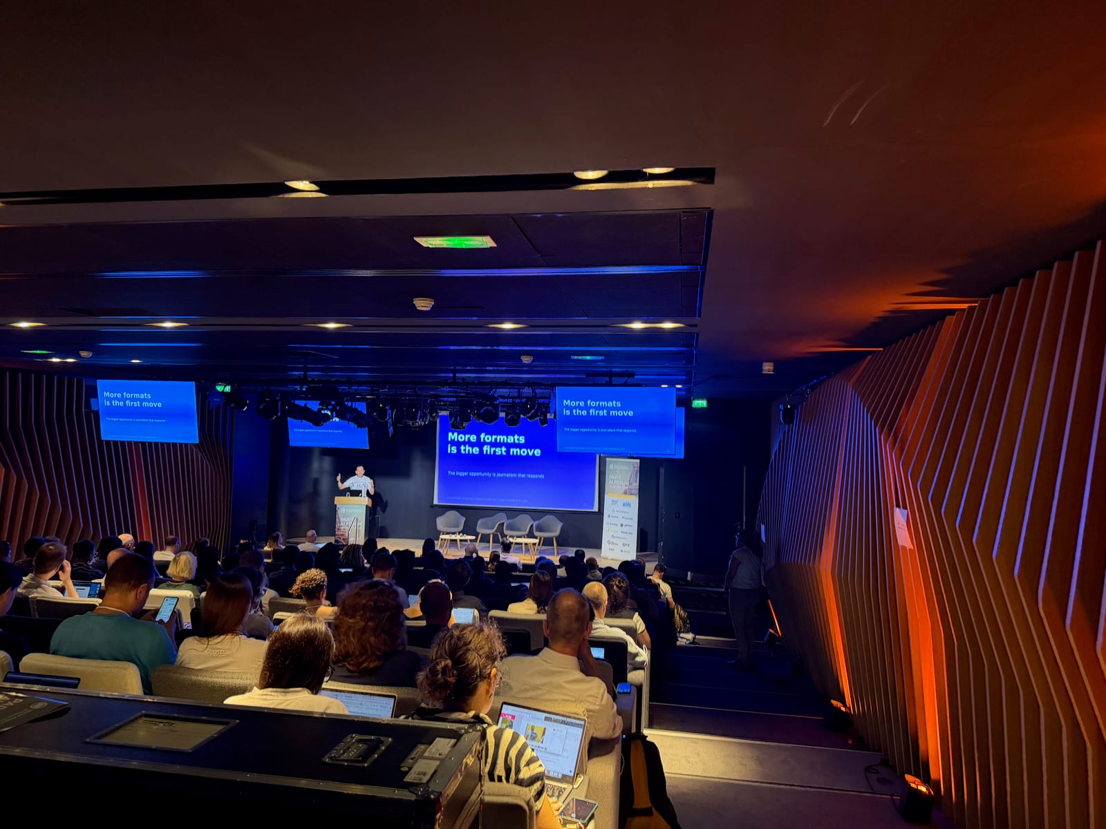 A speaker on stage at WAN-IFRA's Paris event, with slides reading More formats is the first move, facing a full auditorium.